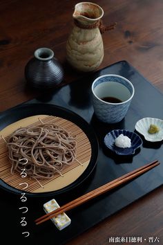 chopsticks and bowls on a black tray with noodles in the middle, two vases behind them