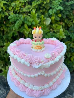 a pink heart shaped birthday cake with a baby doll on top, sitting in front of some bushes