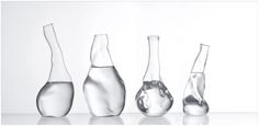 four glass vases are lined up in a row on a reflective surface, with one being filled with water and the other is empty