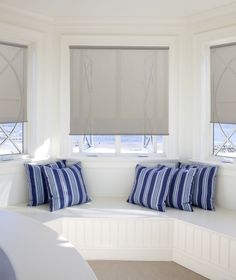 a window seat with pillows on it in front of two windows that have roman shades over them