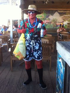 a man in hawaiian clothing holding two bags and a bottle on a wooden deck with people sitting at tables behind him