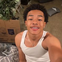 a young man wearing a white tank top sitting in front of a cardboard box and looking at the camera