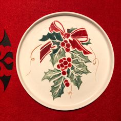 a white plate with holly and red ribbon on it sitting on a red tablecloth