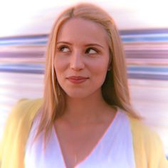 a woman with blonde hair wearing a yellow cardigan and looking at the camera while standing in front of a white wall