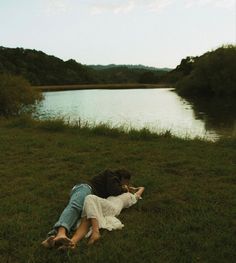 two people laying on the grass next to a body of water with trees in the background