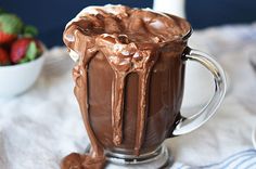 a glass mug filled with chocolate frosting on top of a white table cloth next to a bowl of strawberries