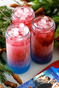 three glasses filled with red, white and blue drink next to pine cones on a table