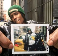 a man holding up a polaroid photo in front of a car on the street