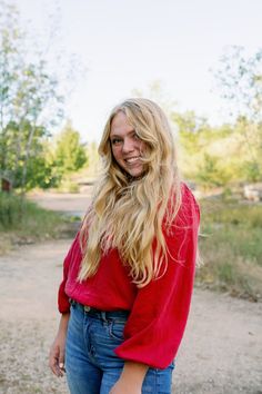 a woman with long blonde hair standing in front of a dirt road and smiling at the camera