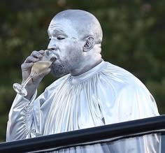 a man with white paint on his face drinking from a wine glass while standing next to a window