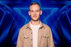 a man standing in front of a blue background wearing a tan jacket and white t - shirt