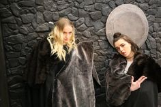 two women standing next to each other in front of a stone wall wearing fur coats