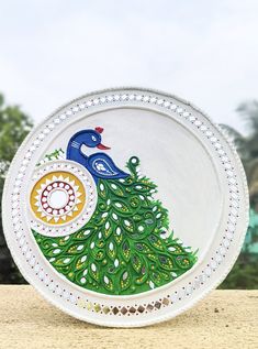 a plate with a peacock painted on it sitting on top of a wooden table next to trees