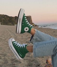 a person sitting on the beach with their feet up and wearing green converse shoes,