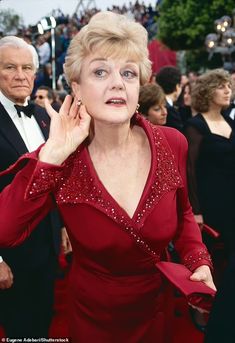 an older woman in a red dress with her hand on her ear and people behind her