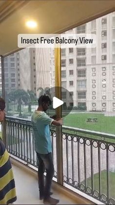 two people standing on a balcony looking out the window at an open area with grass and buildings in the background