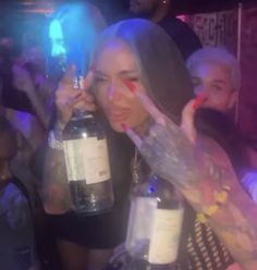 a woman holding up two bottles in front of her face at a party with other people