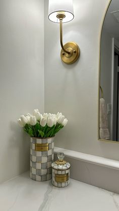 a vase with white flowers sitting on top of a counter next to a mirror and lamp