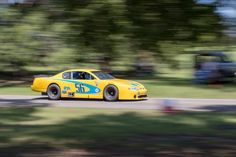 a yellow race car is driving down the road with trees in the backgroud