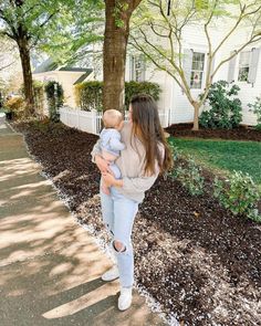 a woman holding a baby in her arms while walking down a sidewalk next to a tree