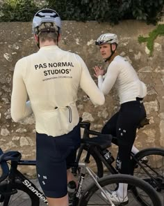 two bicyclists standing next to each other near a stone wall