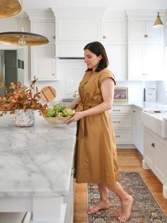 a woman in a brown dress standing at a kitchen island holding a bowl of fruit