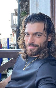 a man with long hair and a beard wearing a black t - shirt sitting on a bench