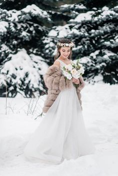 a woman in a white dress and fur coat standing in the snow holding a bouquet