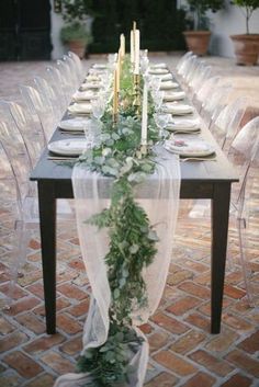 a long table is set with white chairs and greenery on the top, along with candles