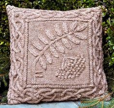 a knitted pillow sitting on top of a wooden table next to some plants and bushes