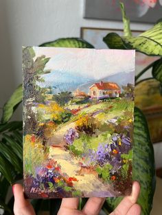 a person holding up a painting in front of a house on a hill with flowers