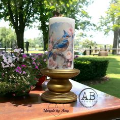 a lit candle sitting on top of a wooden table next to flowers and trees in the background