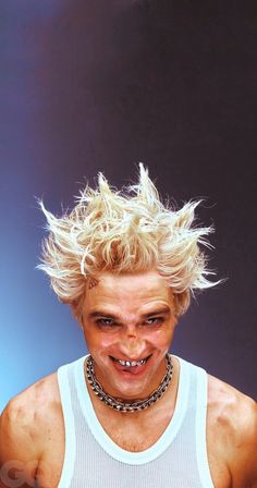 a man with blonde hair and chains on his neck wearing a white tank top is smiling at the camera