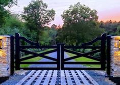 a wooden gate with stone pillars leading to a grassy field at sunset or dawn in the background