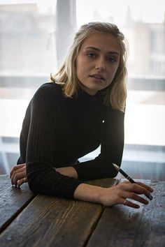 a woman sitting at a wooden table with a pen in her hand and looking off to the side