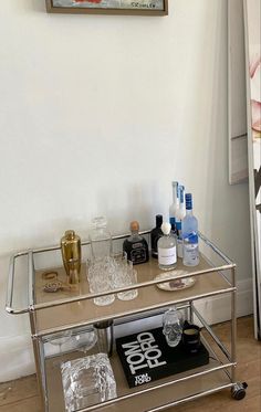 a bar cart with bottles and glasses on it in front of a painting hanging on the wall