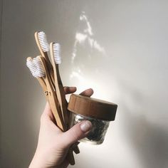 a person holding two wooden toothbrushes and a glass jar filled with toothpaste