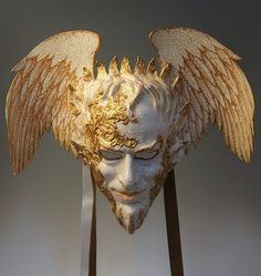 a white mask with gold wings on it's head and two wooden sticks in front of it