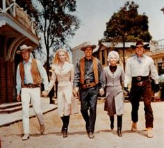 four men and two women are walking down the street in cowboy attire, one is wearing a hat