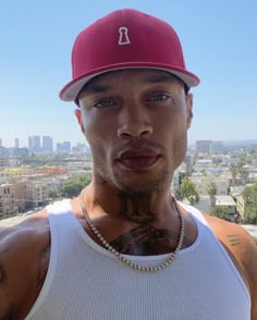 a man with tattoos and a red hat is posing for a photo in front of the city