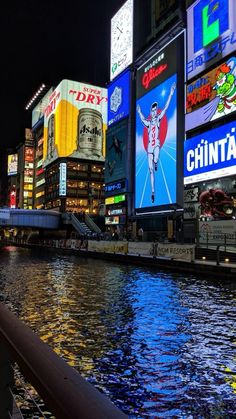 there are many billboards along the water in this city at night, with lights reflecting on the water