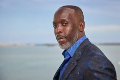 a man in a blue shirt and black blazer looking at the camera while standing by the water