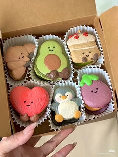 a box filled with assorted decorated cupcakes on top of a table next to a person's hand