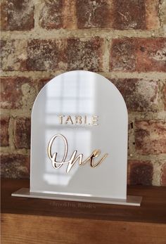 the table one sign is on top of a wooden shelf in front of a brick wall