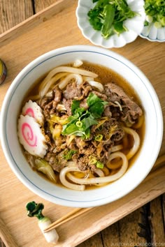 a white bowl filled with noodles and meat on top of a wooden tray next to chopsticks