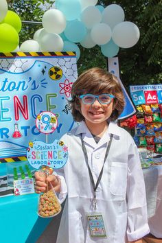 a young boy wearing glasses and holding up a sign at a science fair with balloons in the background