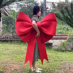 a woman standing in the grass with a large red bow around her neck and hands behind her back