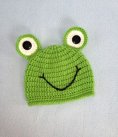 a crocheted frog hat with two eyes and a smile on its face, sitting on a white surface