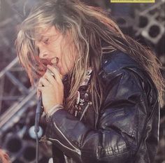 a man with long hair and leather jacket on holding a microphone in his hand while standing next to a stage