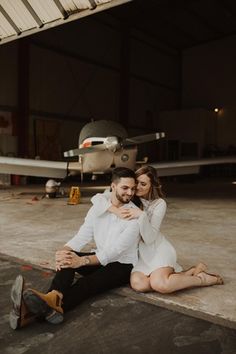 a man and woman are sitting on the ground in front of an airplane with their arms around each other